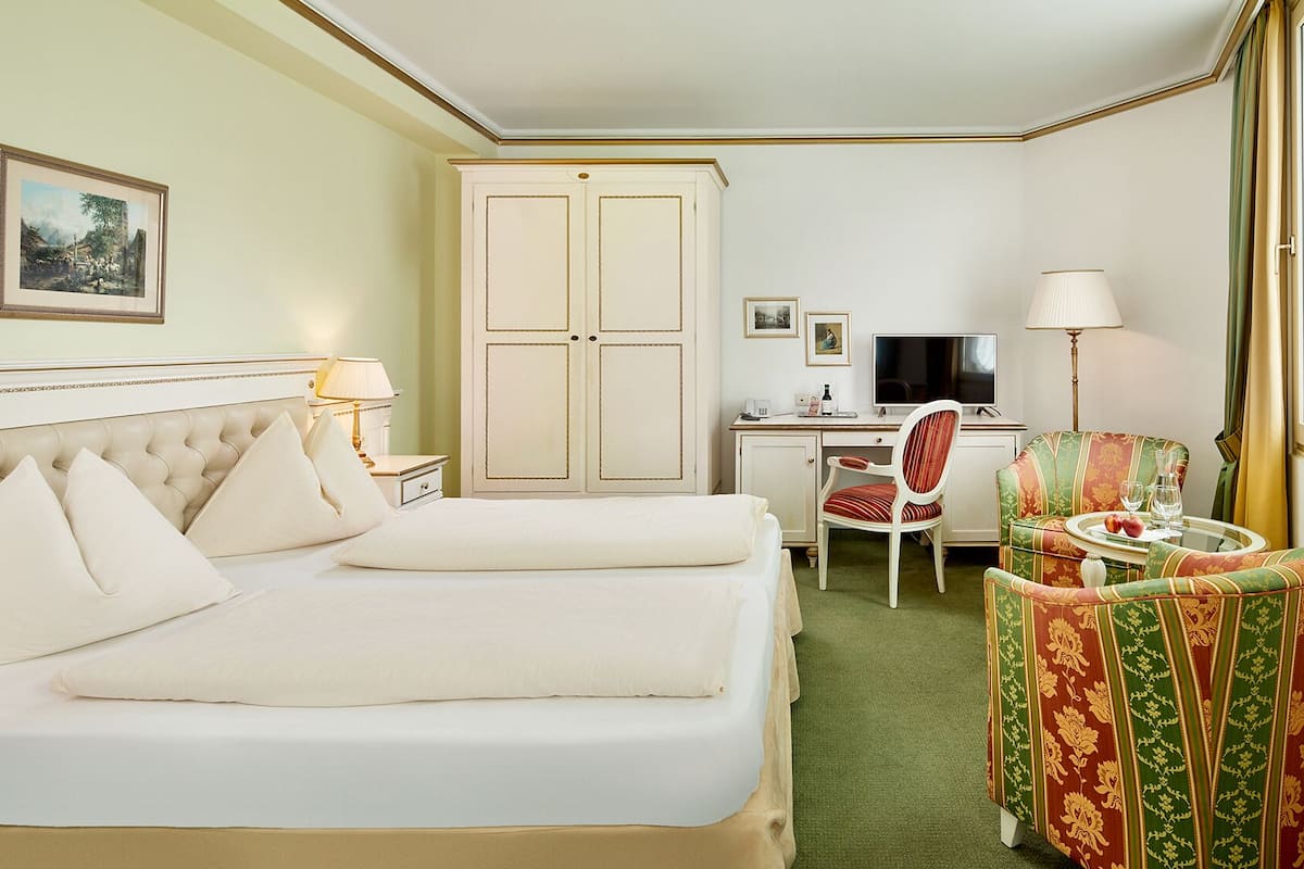 Ein Hotelzimmer mit einem großen Bett, einem Schreibtisch mit Computer, einem Sessel und einem kleinen Tisch mit einem Glas.