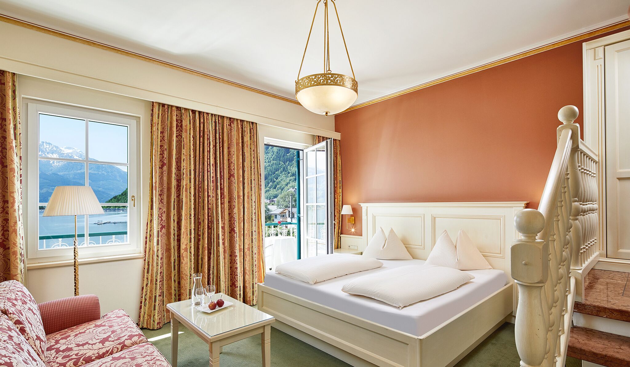 A hotel room with a bed, a small table with a lamp, a view of mountains through the window, and a staircase.