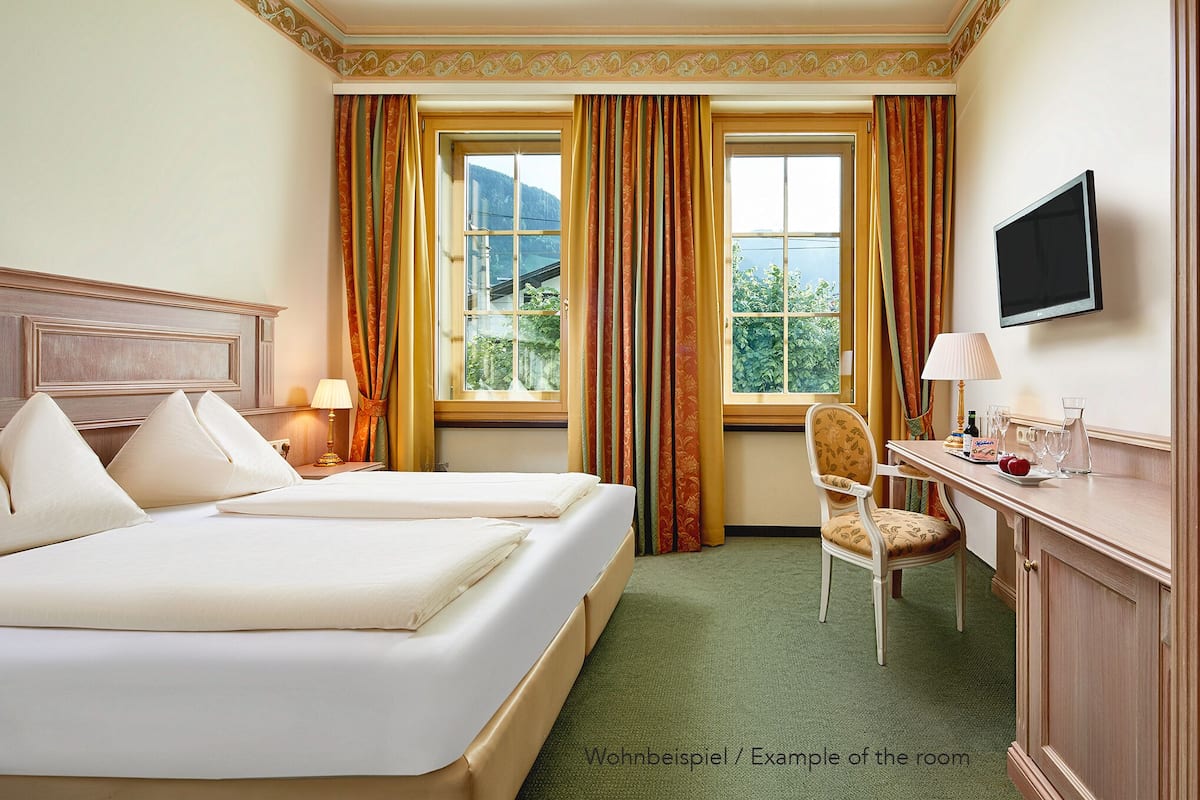 Ein Hotelzimmer mit zwei Betten, einem Schreibtisch, einem Stuhl, einem Fernseher und einem Vorhangfenster.