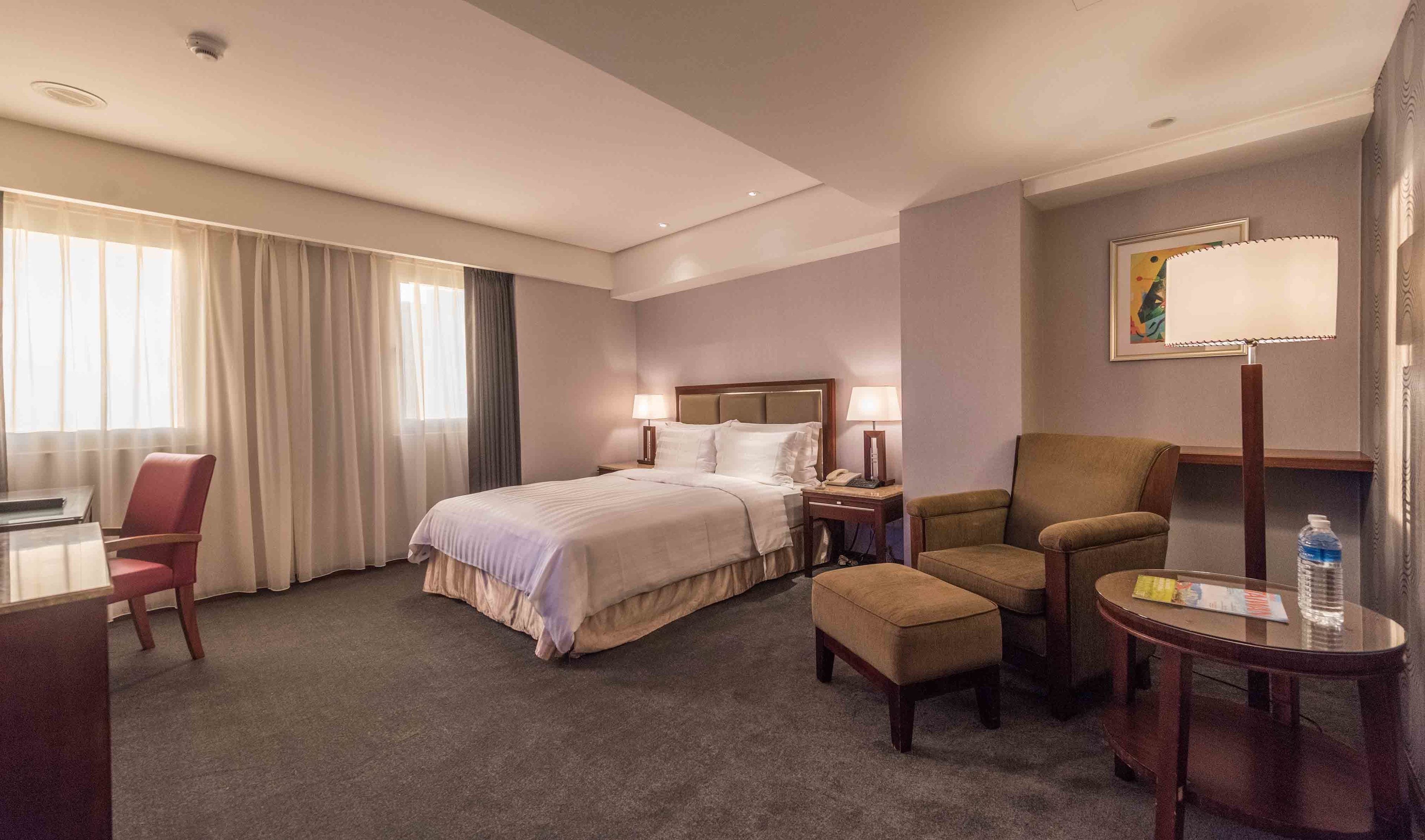 A hotel room with a bed, two chairs, a desk, a lamp, and a painting on the wall.