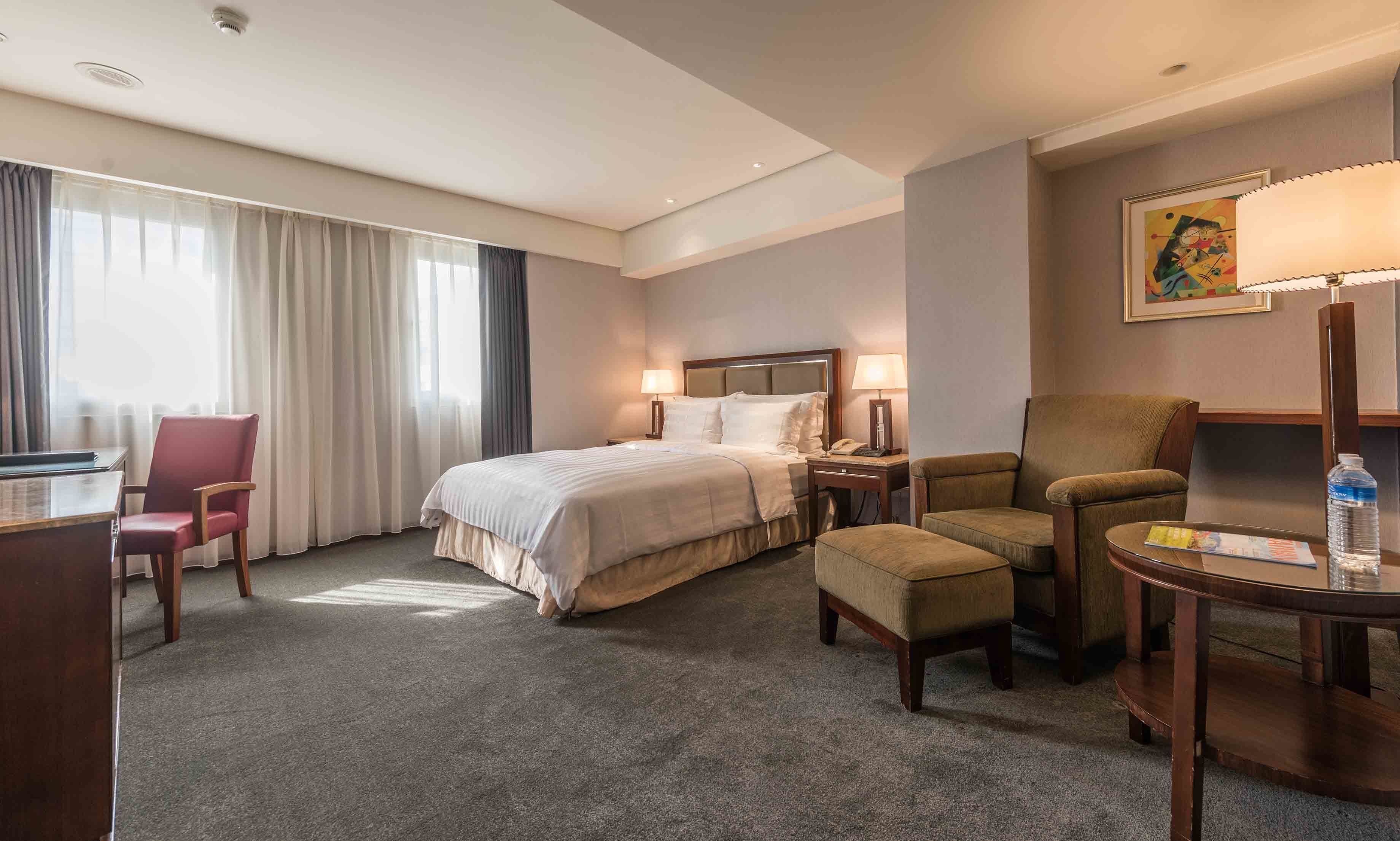 A hotel room with a large bed, two bedside tables with lamps, a desk with a chair, and a small table with a water bottle.