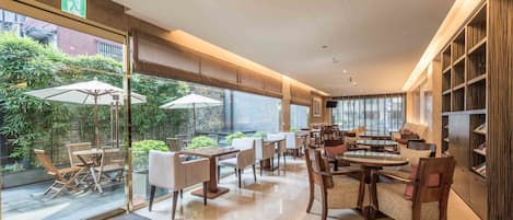 Een moderne hotel lobby met een glazen scheiding, buiten zitplaatsen met parasols, en een grote houten boekenkast.