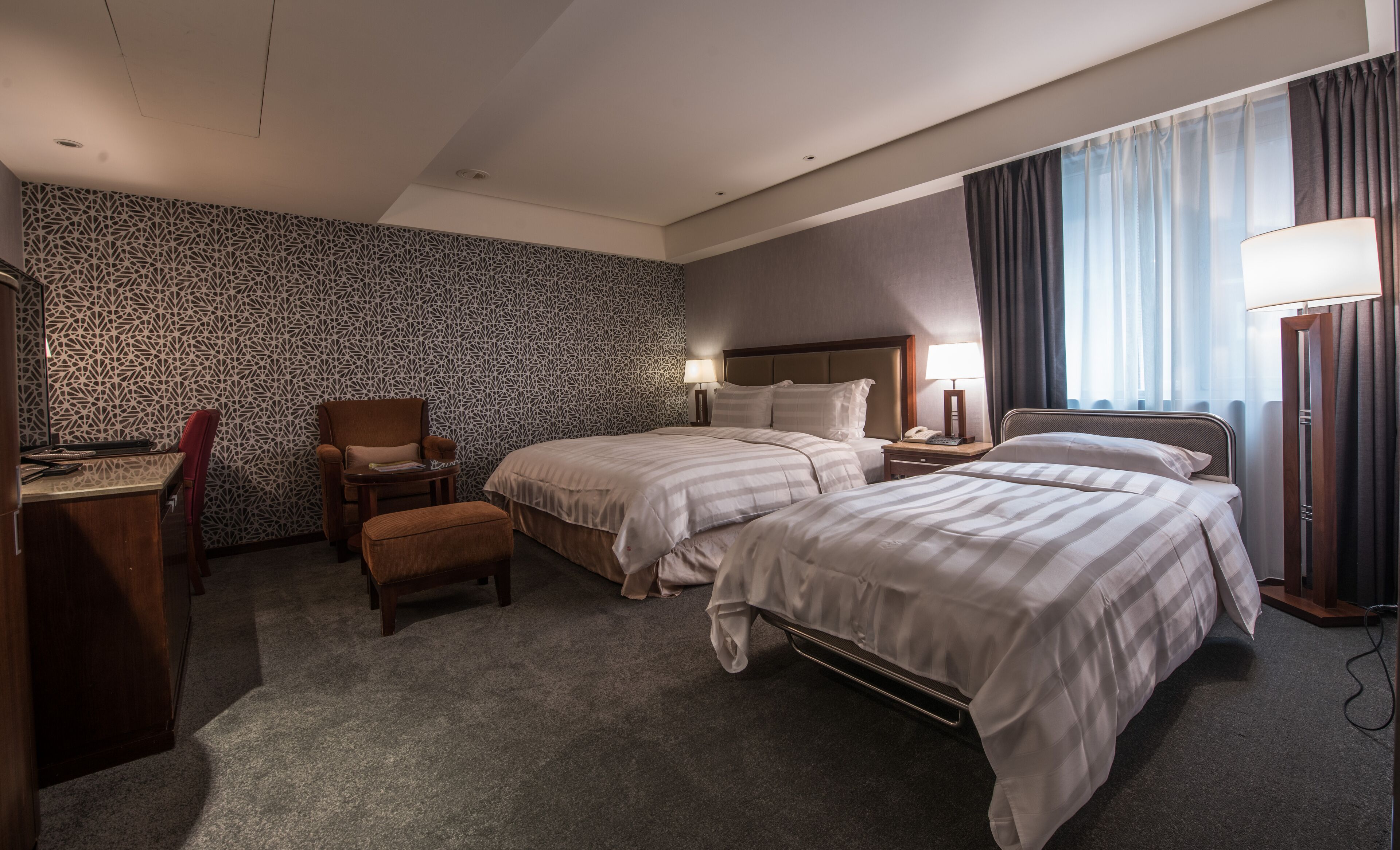 Quarto de hotel com duas camas, uma escrivaninha, uma cadeira e parede estampada.
