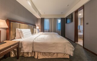 Habitación de hotel con una cama grande, una mesita de noche con lámpara, un televisor montado en la pared y un baño visible a través de una puerta abierta.