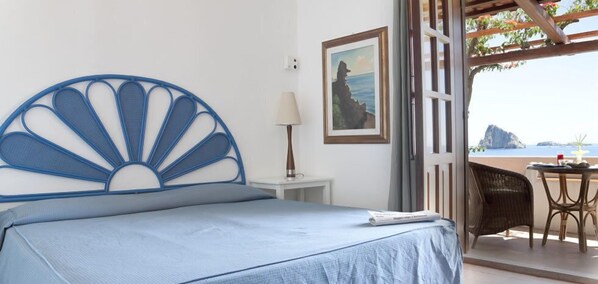 A bed with a blue headboard, a small bedside table with a lamp, a framed picture on the wall, and a view of the sea through an open door. - Hotel La Piazza (Lipari)