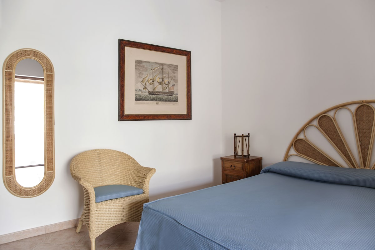 Un dormitorio con una cama, una silla de mimbre con cojín, una mesita de noche de madera, un cuadro en la pared y un espejo grande.