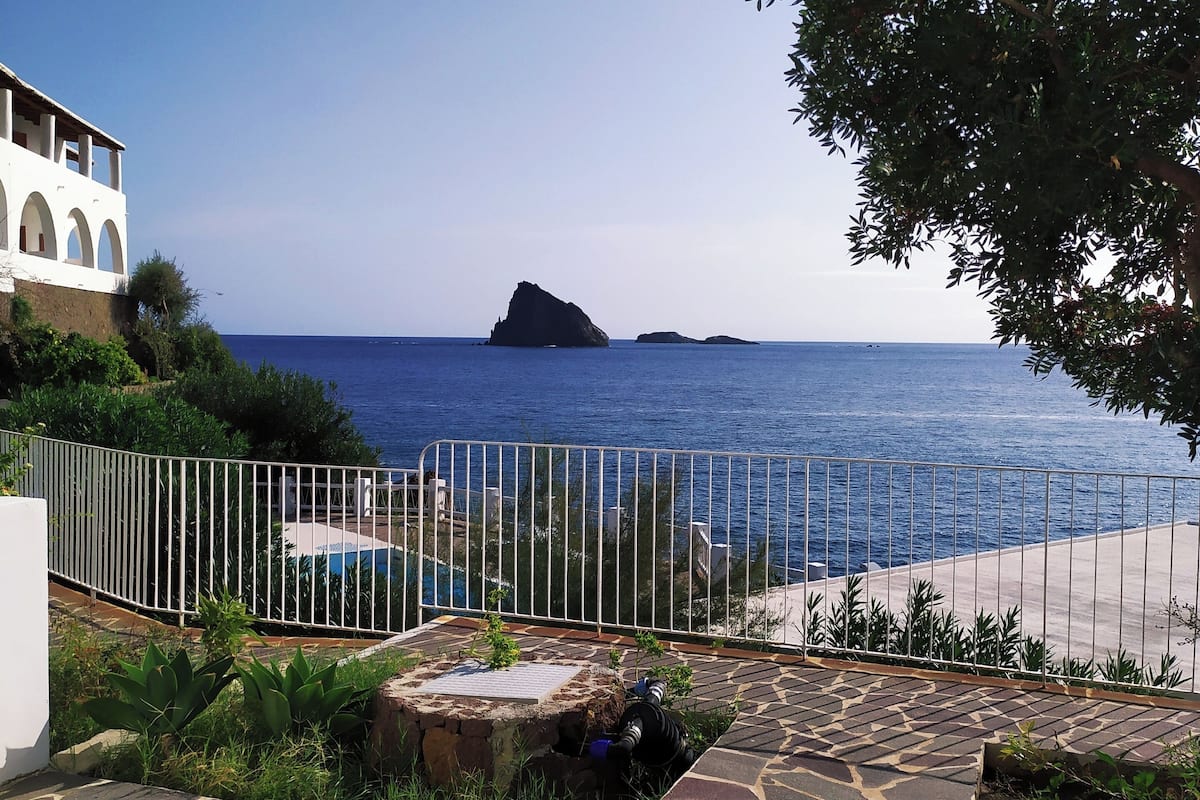 Una propiedad costera con un patio de piedra, una cerca blanca y vistas al mar con una formación rocosa.