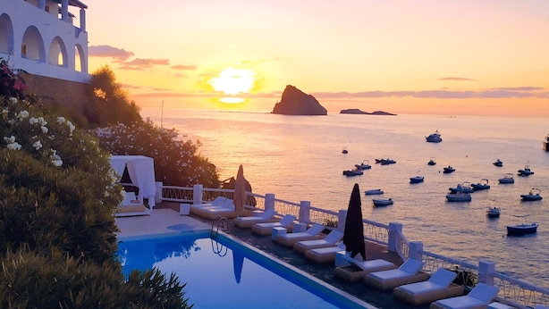 Un área junto a la piscina con sillones y sombrillas, con vistas a un cuerpo de agua con barcos y un atardecer al fondo.