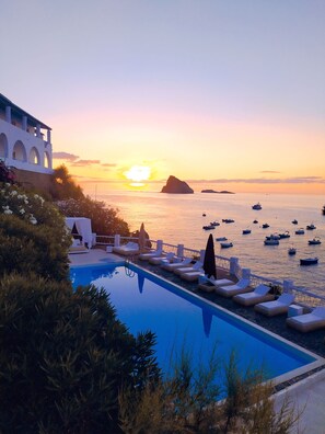 A poolside area with lounge chairs and umbrellas, overlooking a body of water with boats and a sunset in the background. - Hotel La Piazza (Lipari)