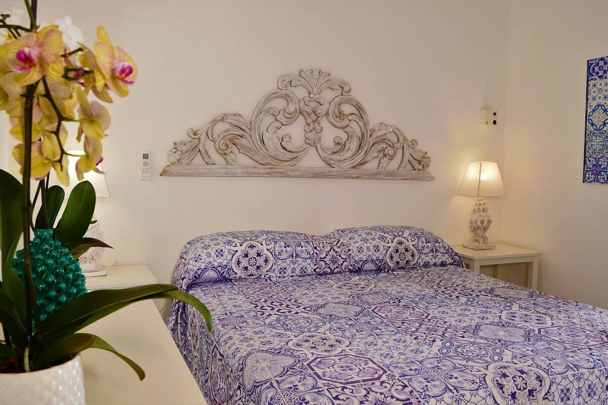 Un dormitorio con una colcha estampada, un cabecero decorativo, una mesita de noche con lámpara y una orquídea en maceta.