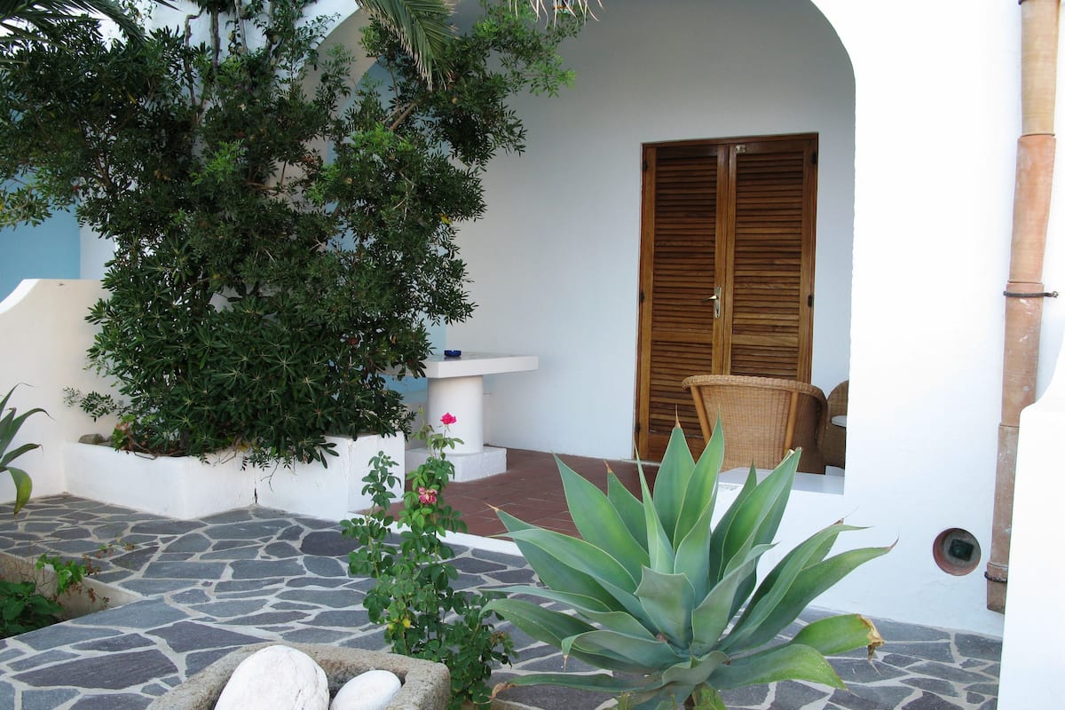 Un patio con suelo de piedra, un banco de piedra, una puerta de madera y una silla de mimbre.