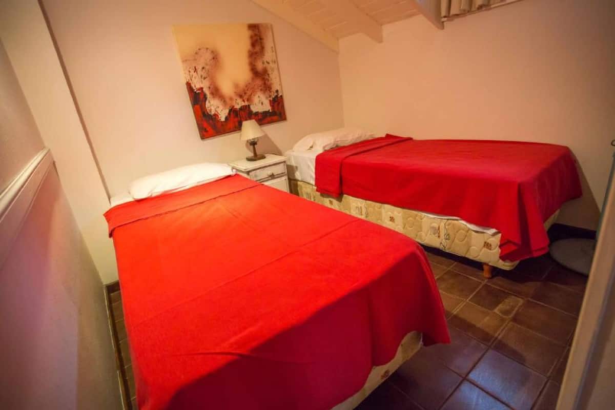 Ein Zimmer mit zwei Betten, jedes mit einer roten Decke bezogen. An der Wand hängt ein Gemälde, zwischen den Betten steht ein Nachttisch mit einer Lampe.