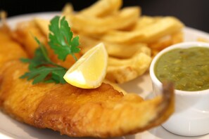 A plate of fish and chips with a lemon wedge and parsley garnish, accompanied by a small bowl of mushy peas.