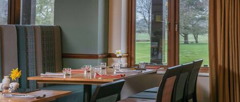 A dining area with a wooden table set for two, cushioned booths, and a view of a grassy landscape through large windows.