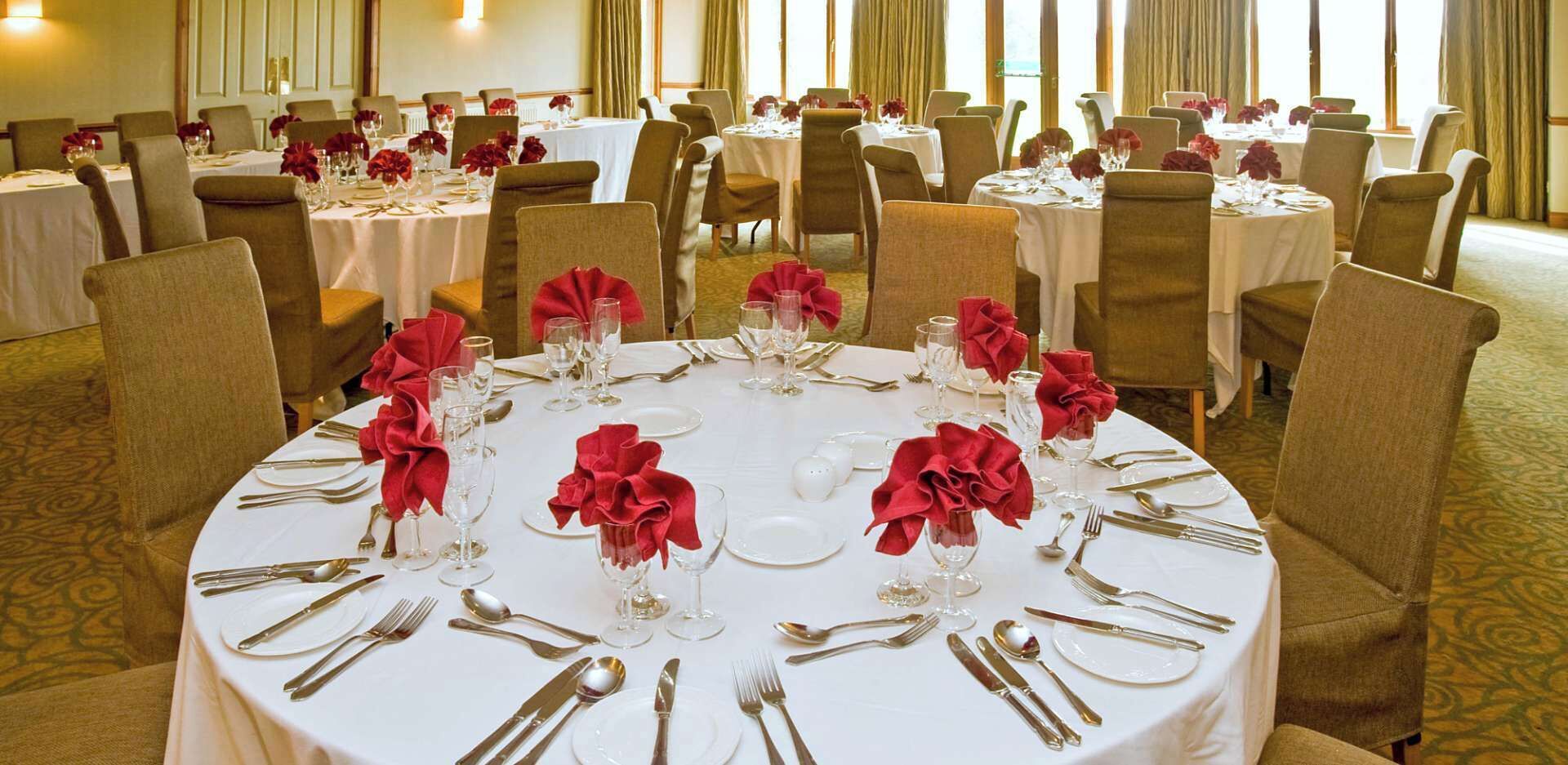 A banquet hall with round tables set for dining, each with white tablecloths, red napkin rings, and silverware.