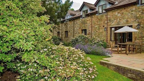 A stone building with a patio area featuring a table and chairs, surrounded by lush greenery and flowering plants. - Garstang Country Hotel & Golf, Sure Hotel Collection (Preston)
