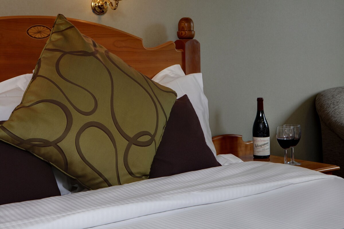 Un lit bien fait, avec une tête de lit en bois, une bouteille de vin et un verre de vin rouge posés sur une table de chevet.