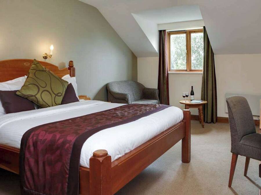 A hotel room with a large bed, a desk with a chair, a small table with a bottle and glasses, and a window with curtains.