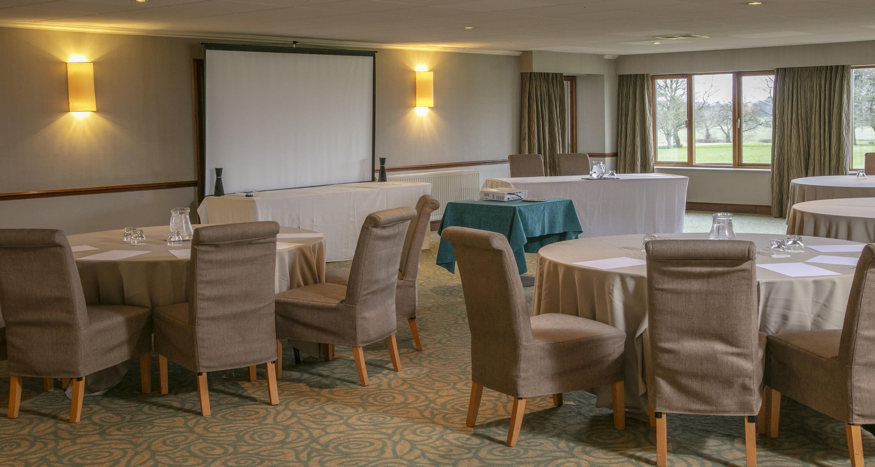A conference room with round tables, beige chairs, and a projector screen.