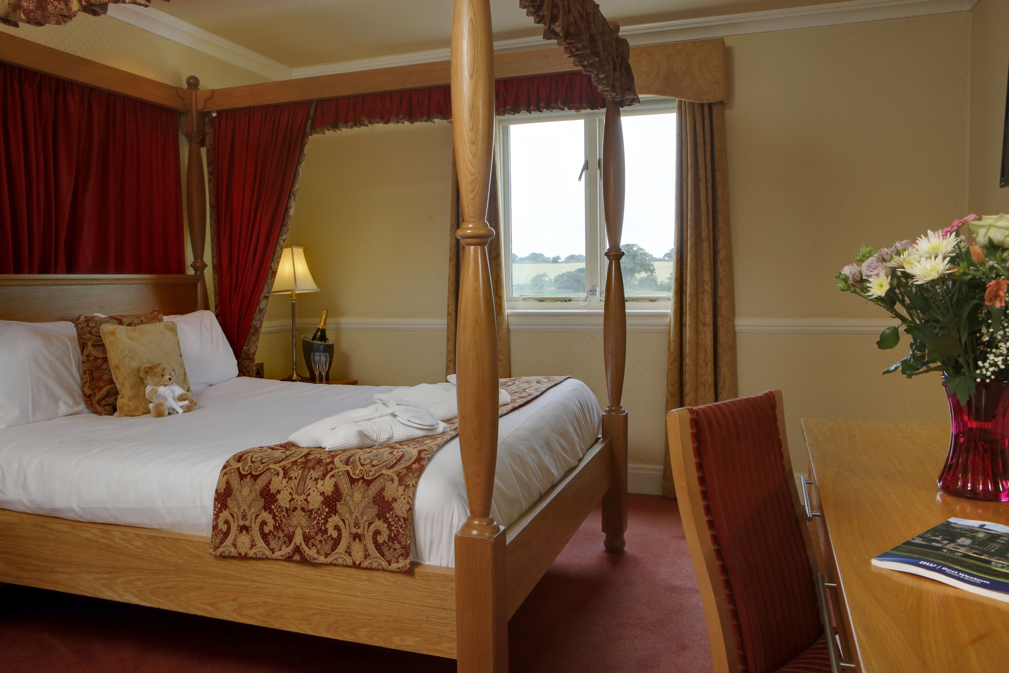 A four-poster bed with a wooden frame, a red and gold bedspread, and a teddy bear on the pillows. There is a wooden table with a chair and a vase of flowers.