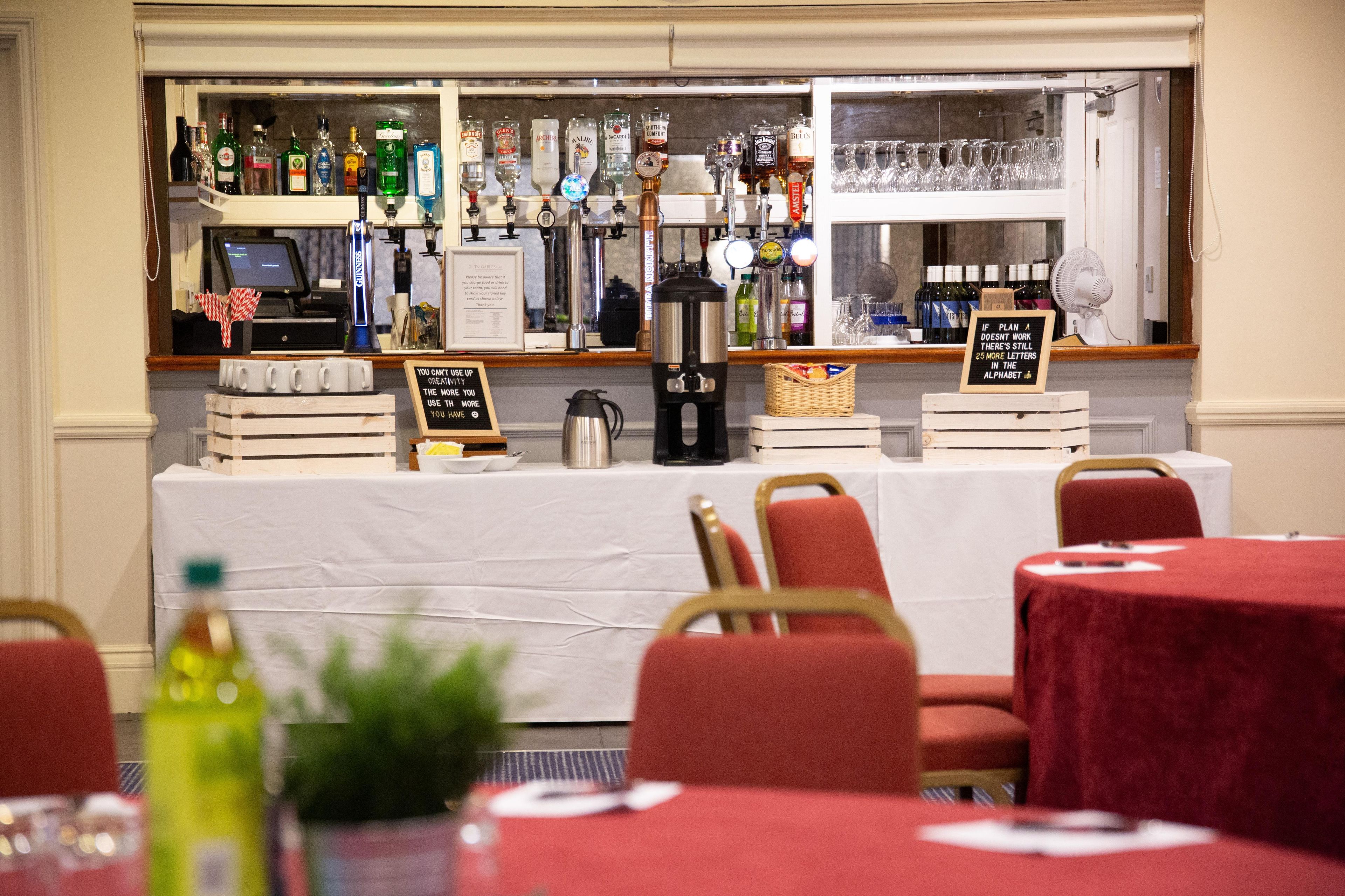 Un coin bar avec différentes bouteilles d’alcool, un appareil à café et un panneau affichant un message. Des tables et des chaises sont disposées au premier plan.