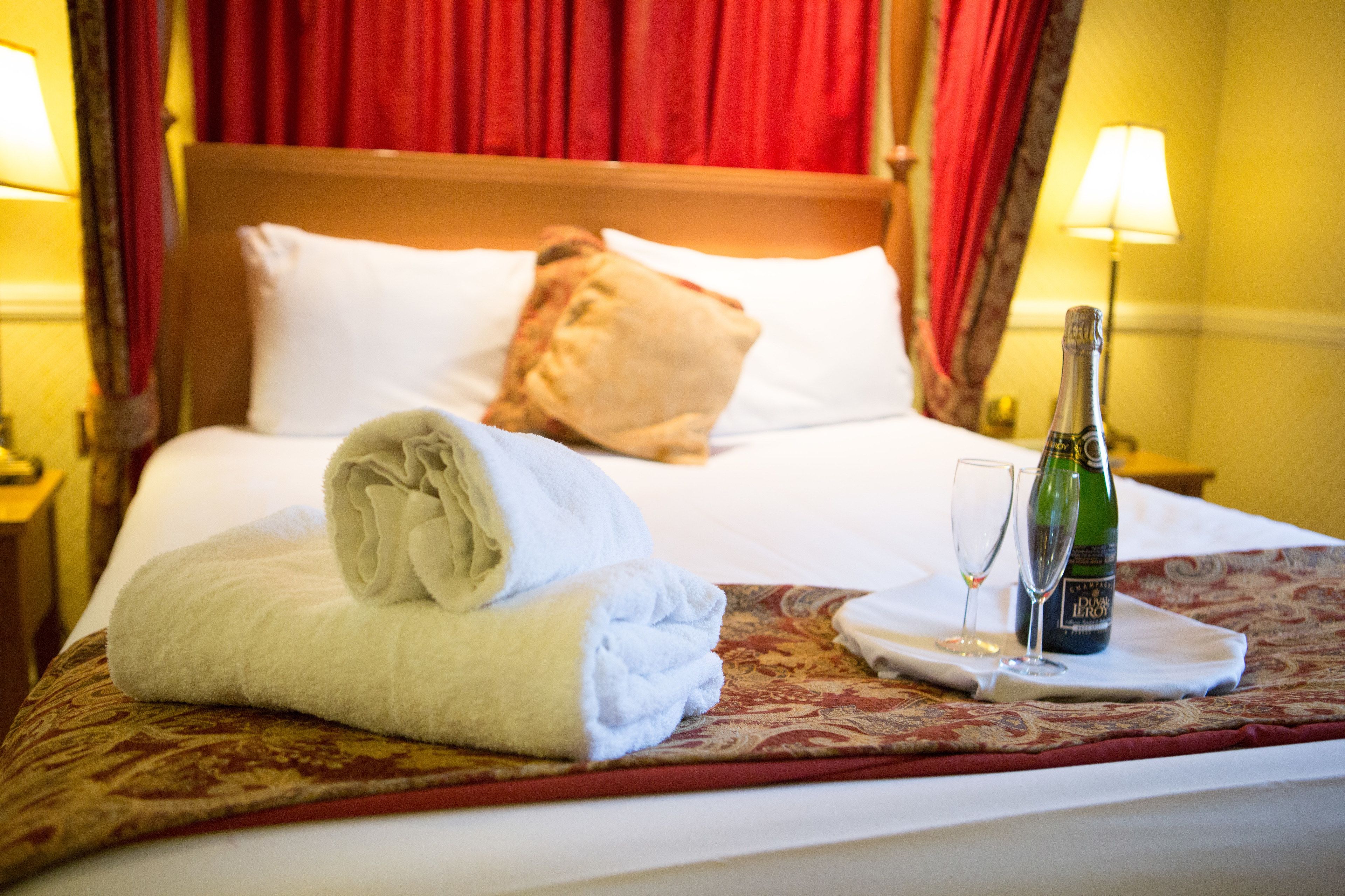 A neatly made bed with a folded white towel, a bottle of champagne, and two glasses on a tray.