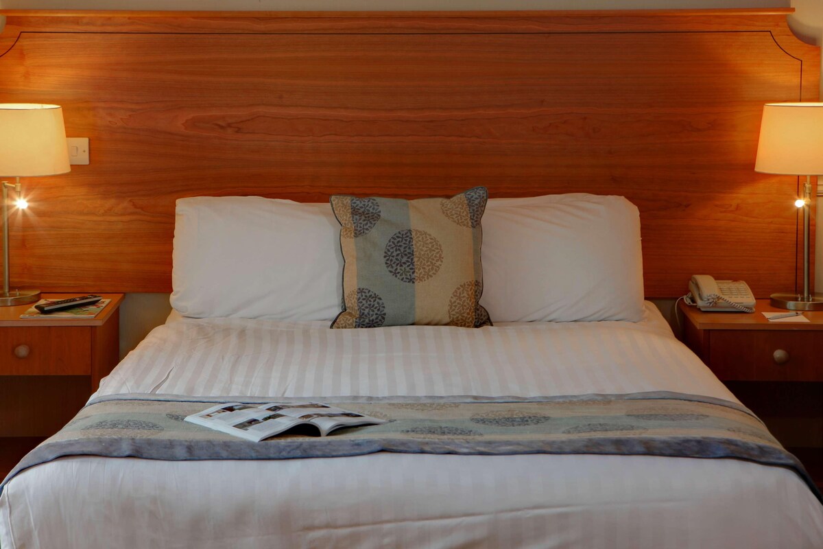 A hotel room with a wooden headboard, two bedside lamps, a telephone, and a magazine on the bed.