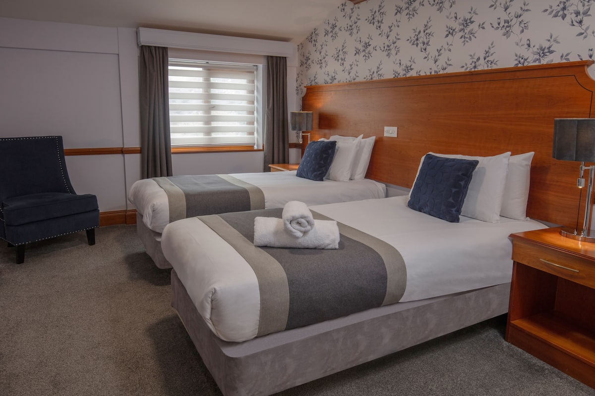 Una habitación de hotel con dos camas, una silla, una mesita de noche y una ventana con cortinas.
