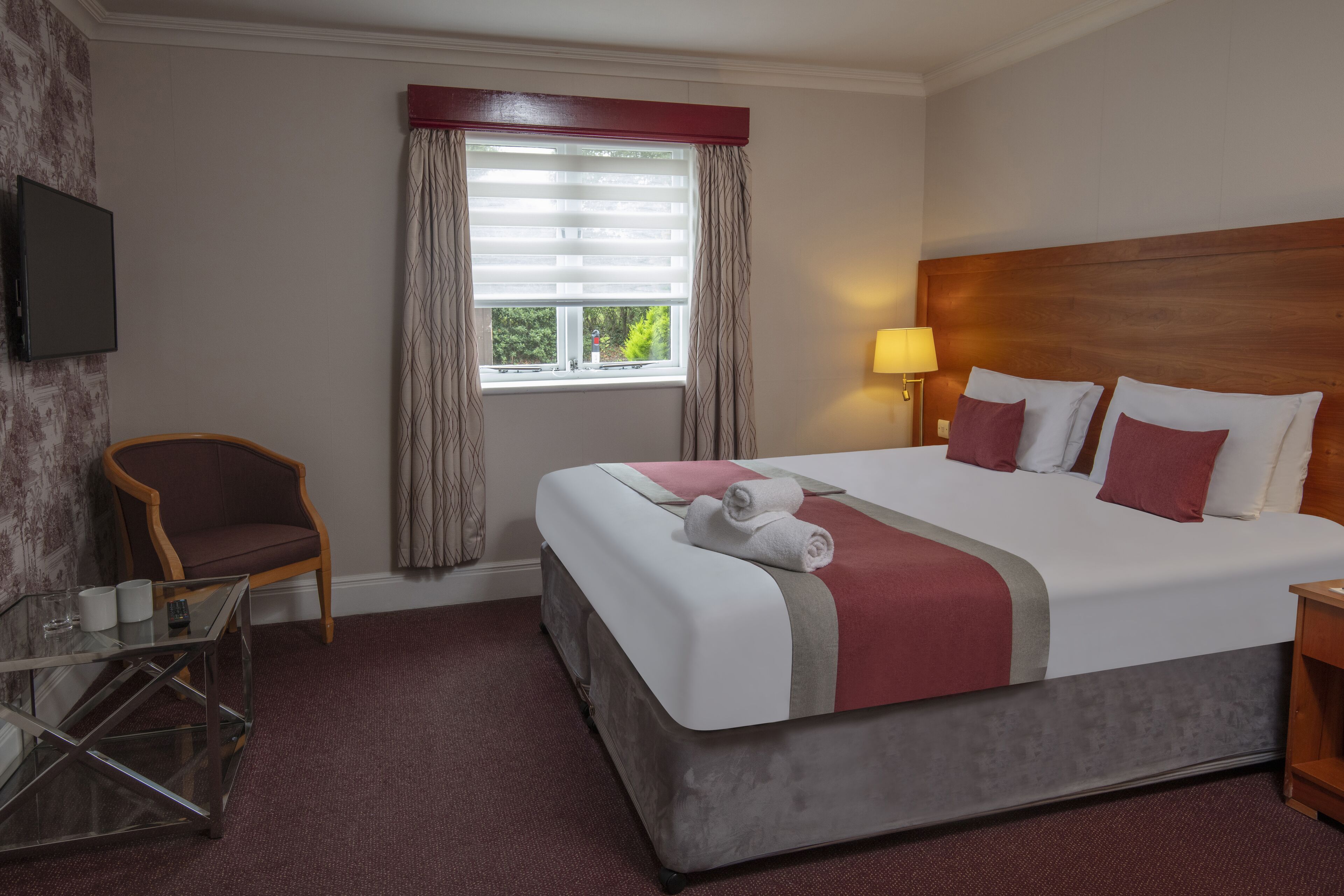Een hotelkamer met een groot bed, een aan de muur bevestigde televisie, een stoel, een glazen tafel en een raam met gordijnen.