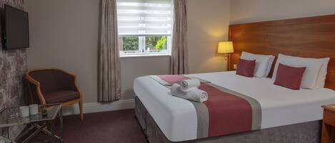 Een hotelkamer met een groot bed, een aan de muur bevestigde televisie, een stoel, een glazen tafel en een raam met gordijnen.
