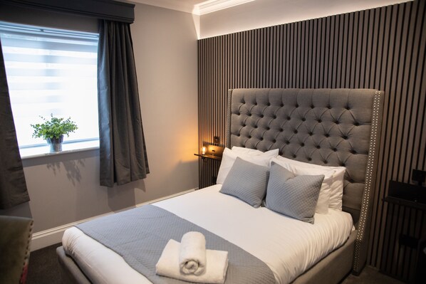 A neatly made bed with a grey headboard, a bedside lamp, and a window with curtains. - Best Western Bristol North The Gables Hotel (Wotton-under-Edge)