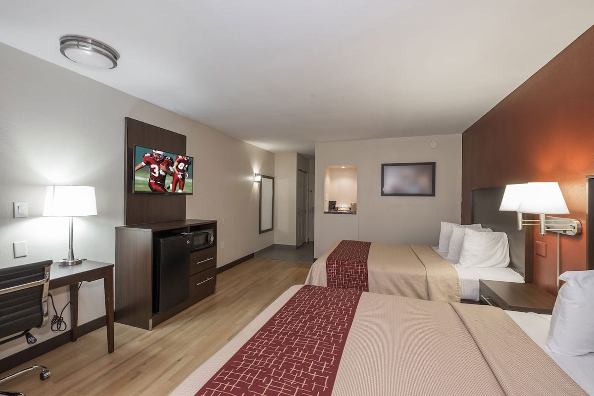 Quarto de hotel com duas camas, uma escrivaninha com luminária, televisão e minibar.