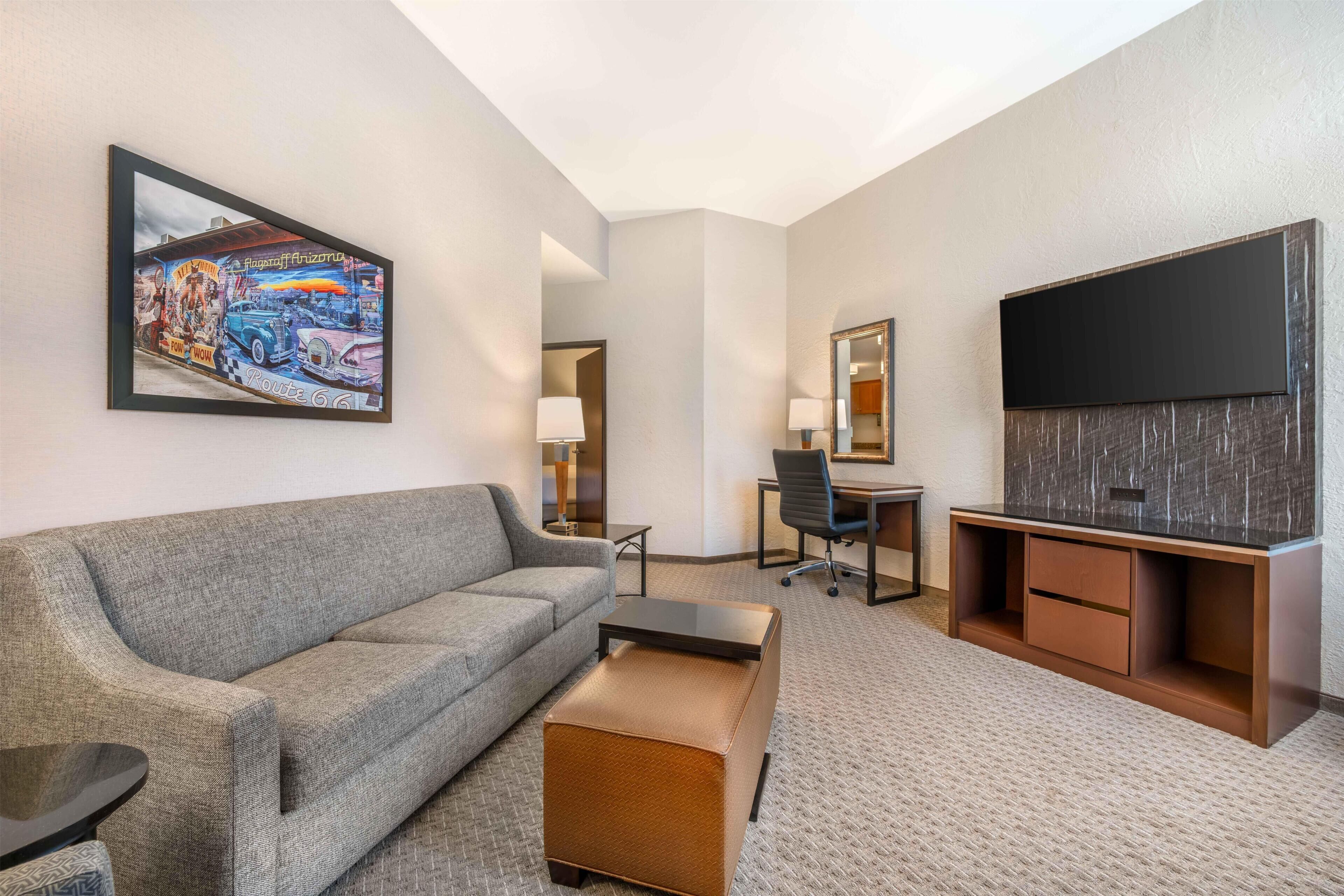 Ein Hotelzimmer mit Sofa, Hocker, Schreibtisch, Stuhl, Fernseher und einem Kunstwerk an der Wand.