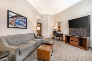 A hotel room with a sofa, ottoman, desk, chair, TV, and artwork on the wall. - Drury Plaza Hotel Flagstaff (Flagstaff)