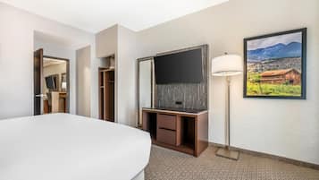 A hotel room with a large bed, a flat-screen TV mounted on a wooden cabinet, a desk with drawers, a standing lamp, and a framed picture of a rural landscape on the wall.