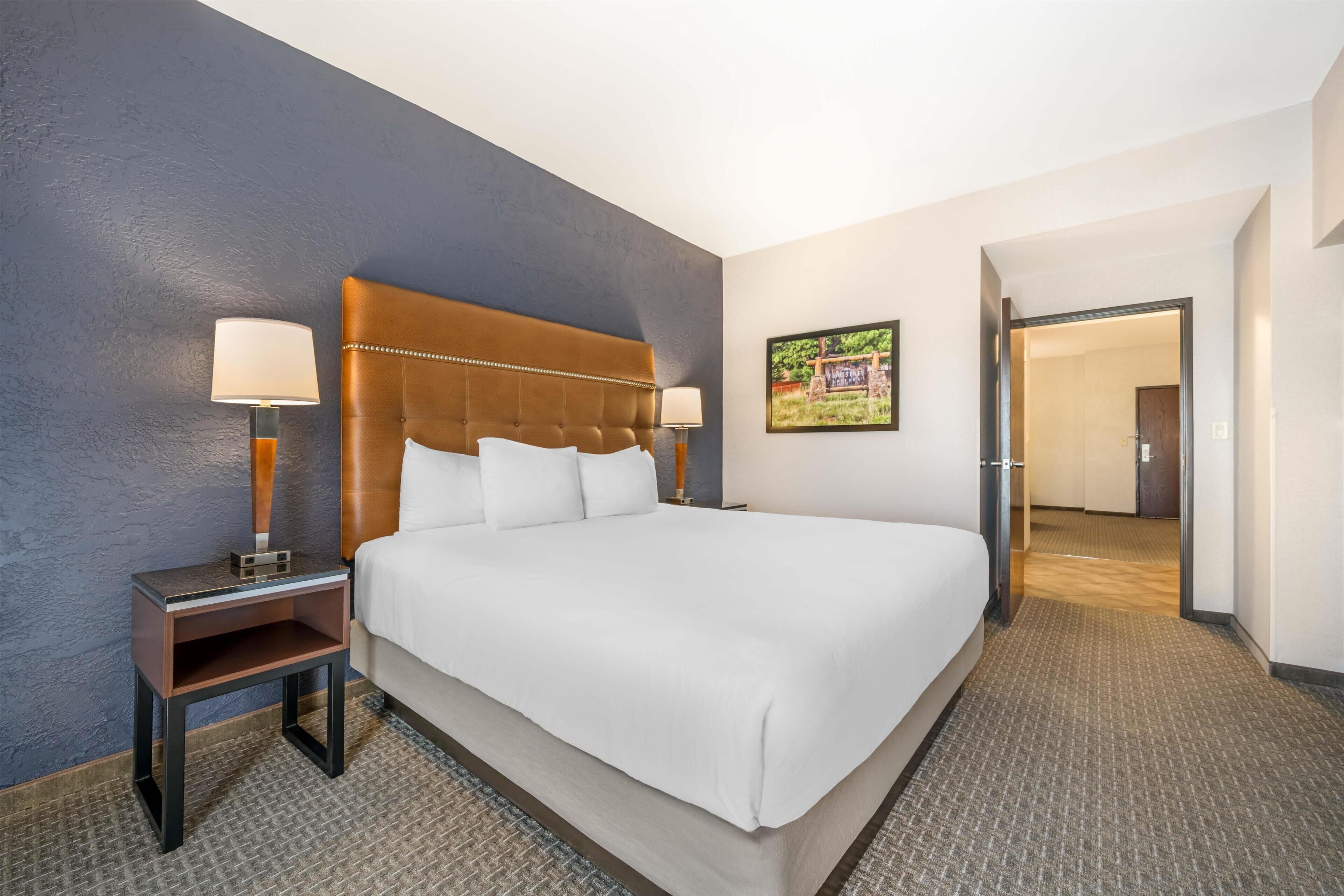 A hotel room with a large bed, two bedside tables with lamps, a framed picture on the wall, and a door leading to another room.