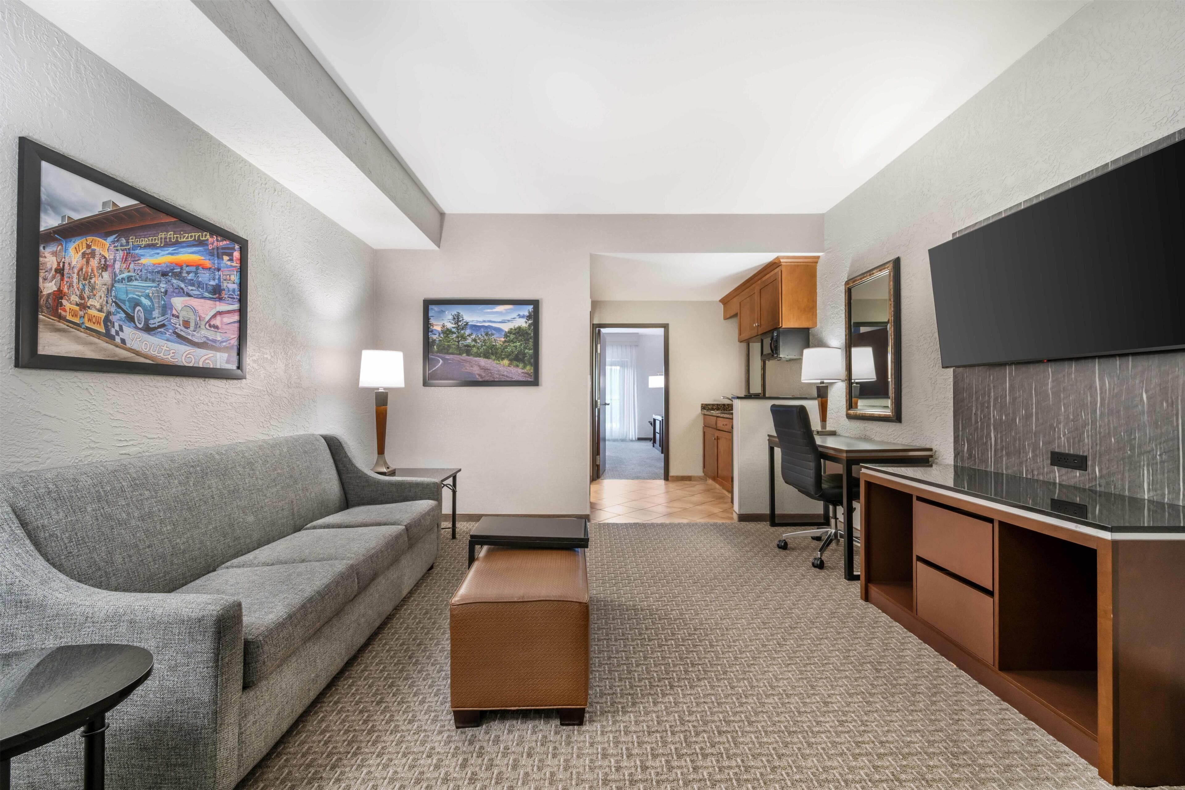 A hotel room with a gray sofa, a small table, a TV, a desk, and a cabinet. There is a painting on the wall and a door leading to the hallway.