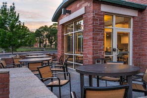 An outdoor seating area with tables and chairs, a brick building with large windows, and a view of trees and a road in the background. - Drury Plaza Hotel Flagstaff (Flagstaff)