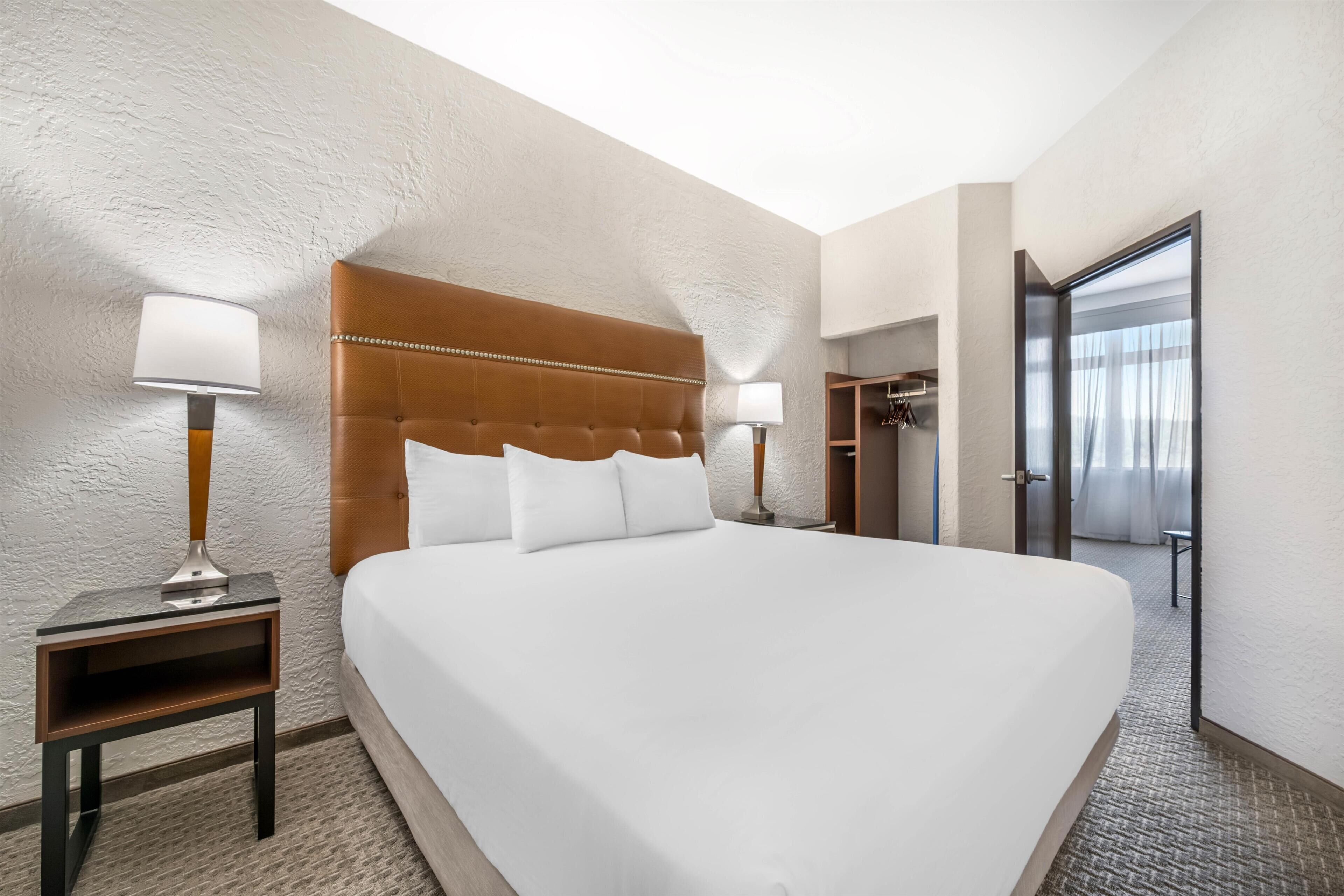 A hotel room with a large bed, two bedside tables with lamps, a wooden headboard, and a door leading to a bathroom.