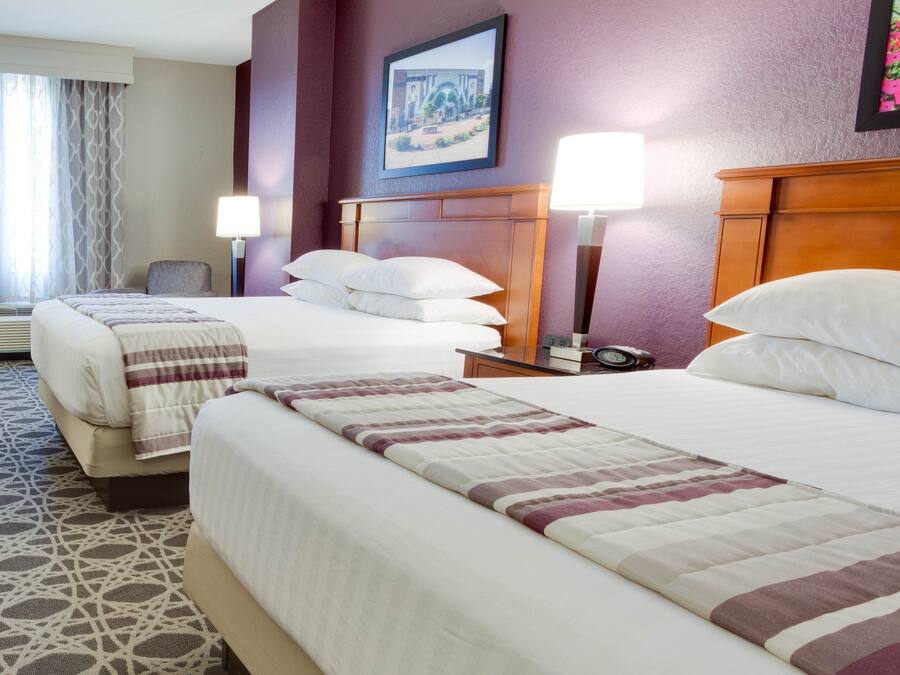A hotel room with two beds, a nightstand, a lamp, a clock, and a framed picture on the wall.