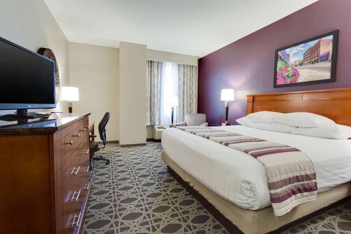 A hotel room with a large bed, a desk with a flat-screen TV, two bedside lamps, and a window with curtains.