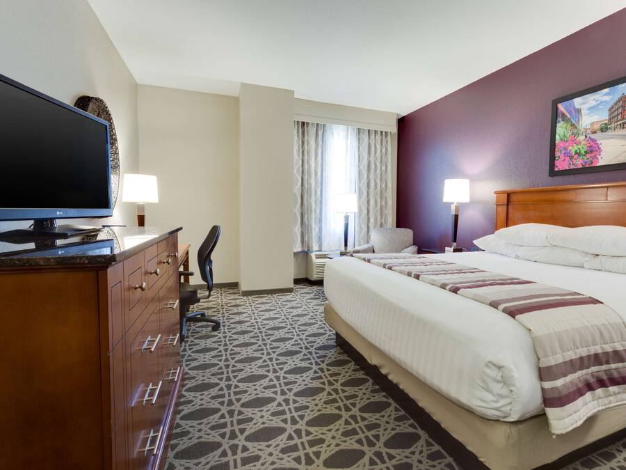 Ein Hotelzimmer mit einem großen Bett, einem Schreibtisch mit Flachbildfernseher, zwei Nachttischlampen und einem fensterbelaubten Fenster.
