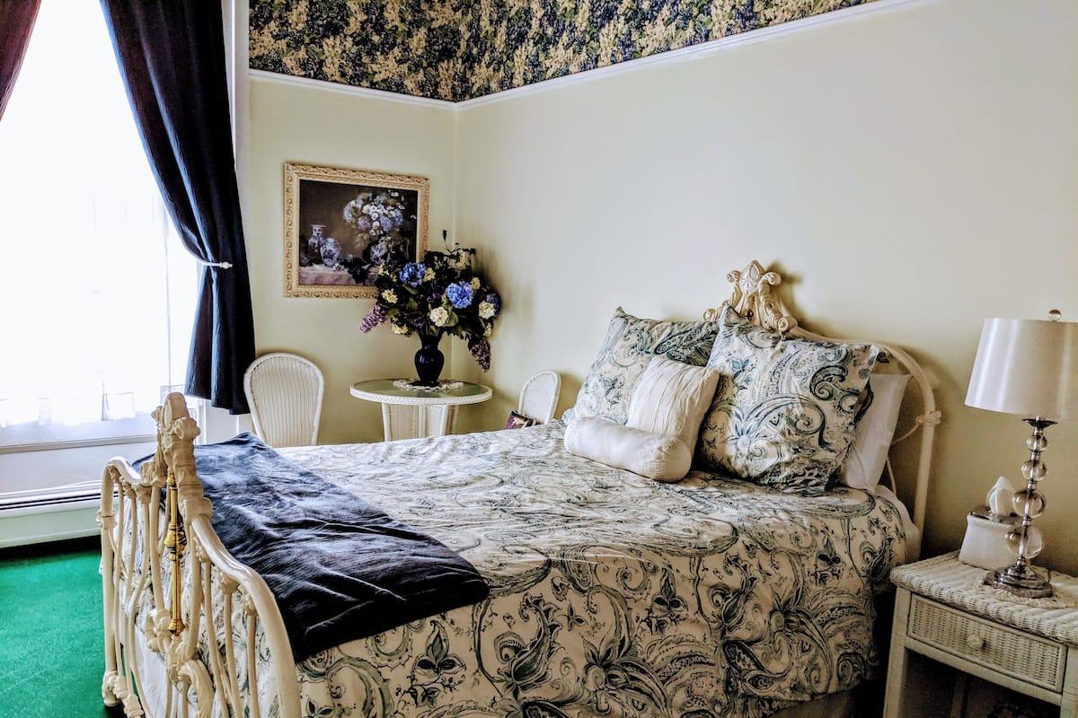 A bedroom with a bed, a bedside table, a chair, a window with curtains, and a painting on the wall.