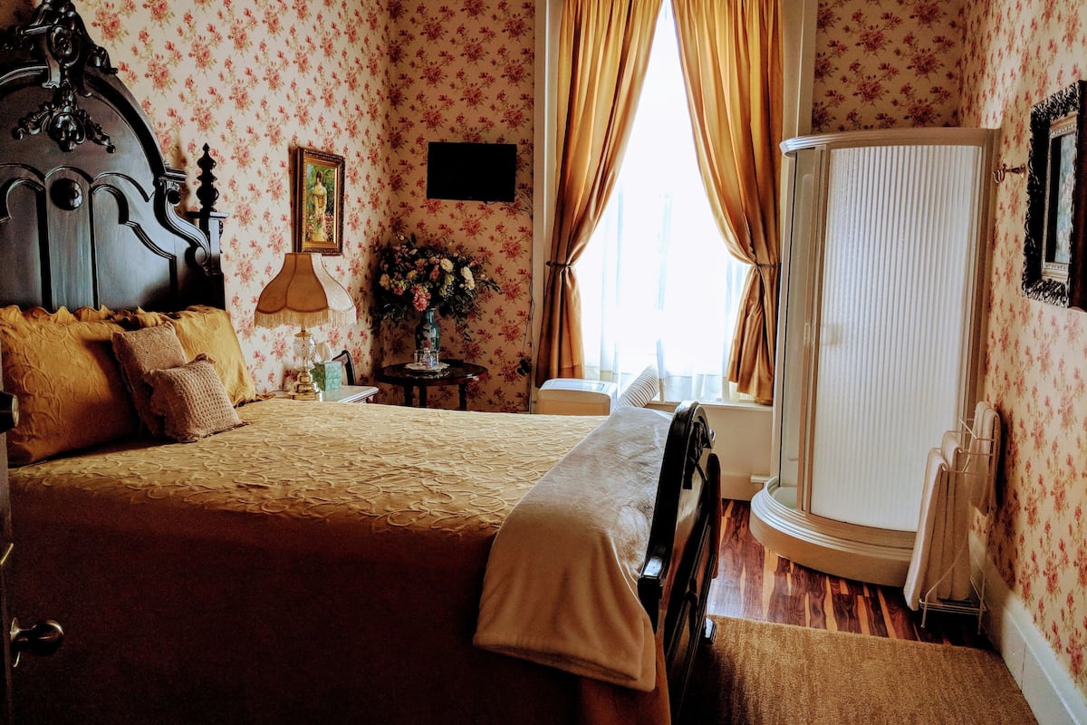 A bedroom with a large bed, a nightstand, a dresser, a window with curtains, and a mirror.