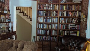 A library with a large bookshelf filled with books, a leather armchair, and a patterned sofa. - Allegiance Bed and Breakfast (Mount Morris)