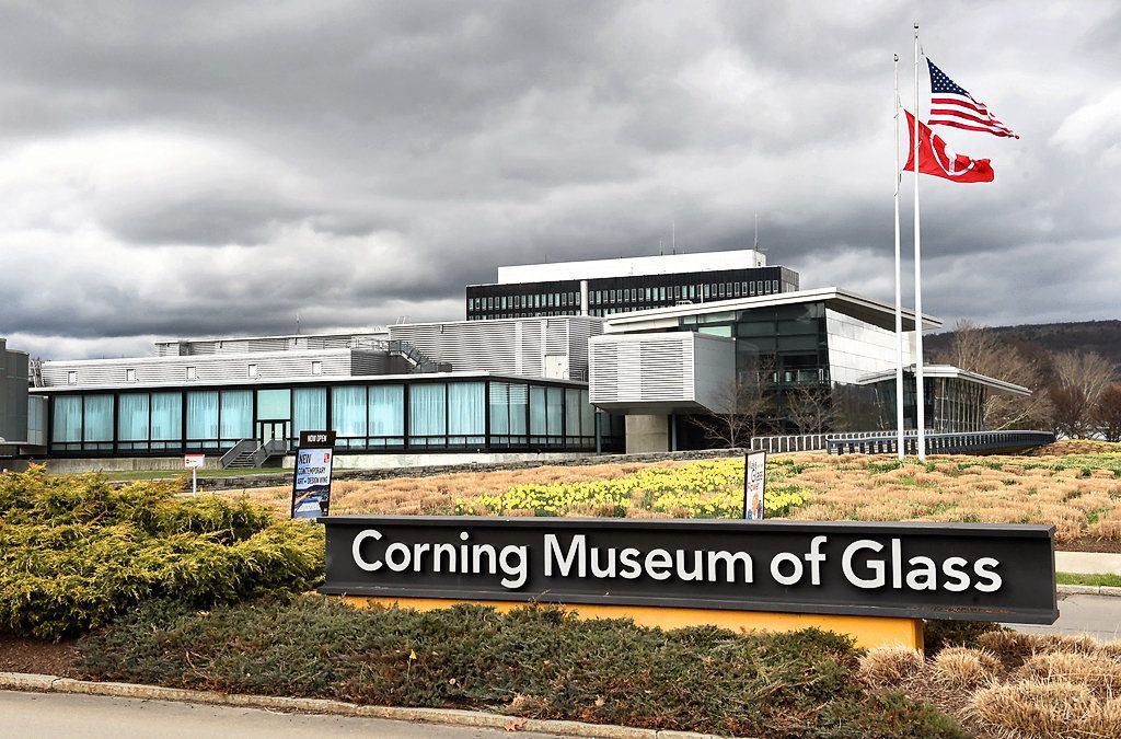 The Corning Museum of Glass building with a signboard, American flag, and surrounding landscaping.
