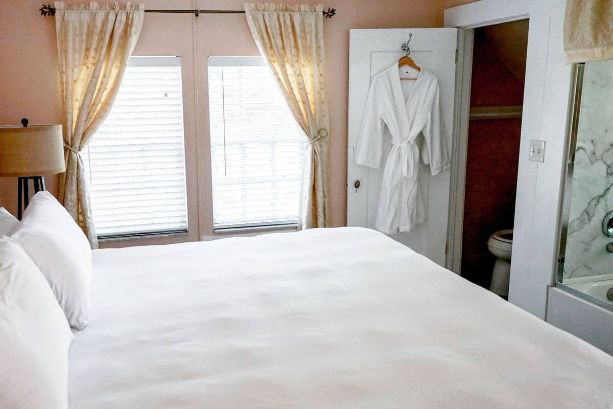 Ein Hotelzimmer mit einem großen Bett, zwei Fenstern mit Vorhängen, einer Nachttischlampe, einem Bademantel, der an einer Tür hängt, und einem Badezimmer mit Marmor.