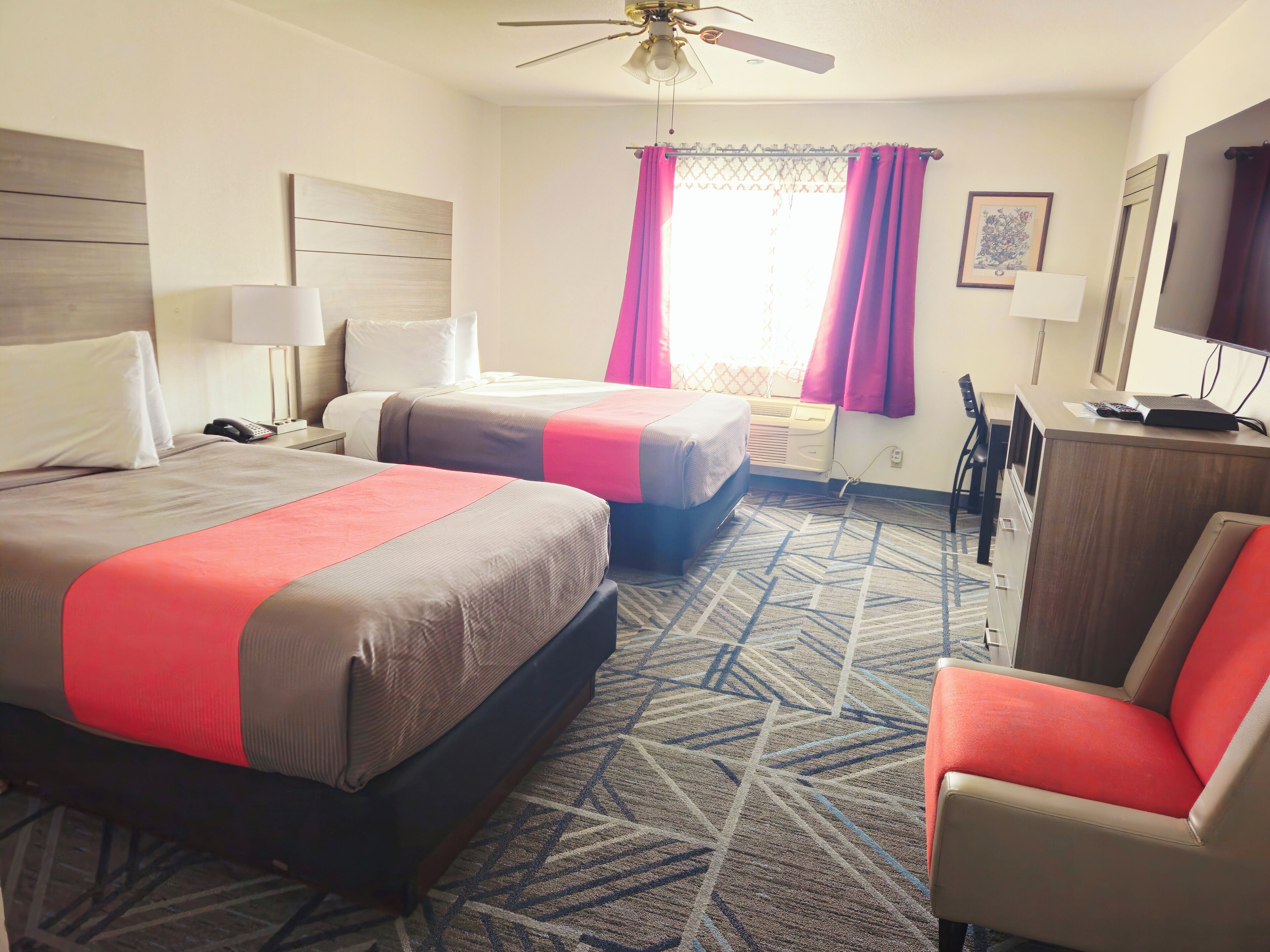 A hotel room with two beds, a desk, a chair, a ceiling fan, and a window with curtains.