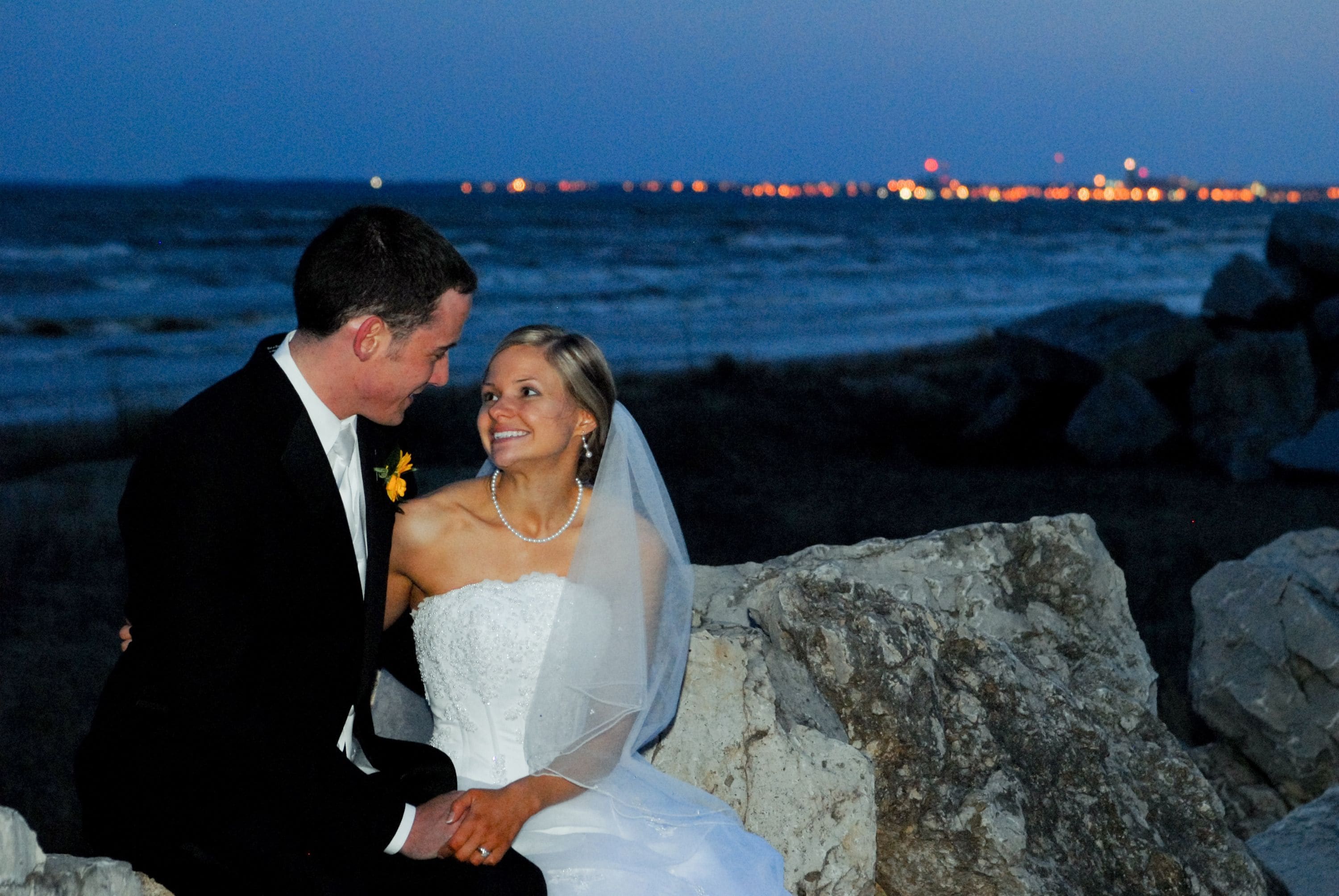 A couple seated on rocks by the ocean at night with city lights in the background.