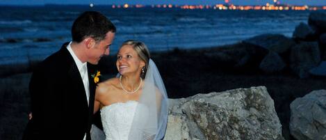 A couple seated on rocks by the ocean at night with city lights in the background.